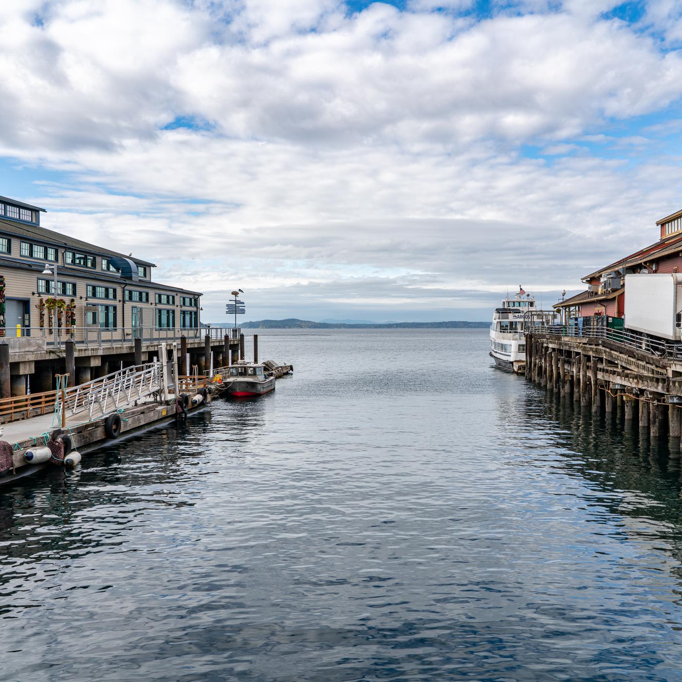Calm water framed by two wooden piers on the Seattle waterfront, with a small work boat at left and Puget Sound stretching to distant hills